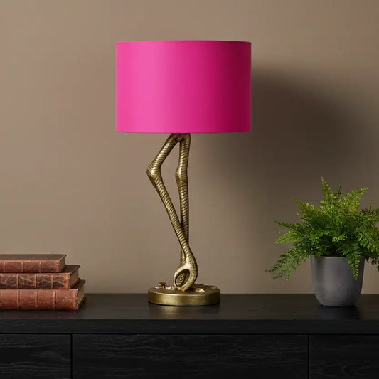 Table lamp with pink shade and gold base on a dark surface against a brown wall.