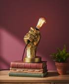 Gold fist lamp with an exposed bulb on stacked books against a purple wall.