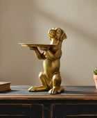 Gold dog statue holding a tray on a wooden surface with a plant and book in the background.