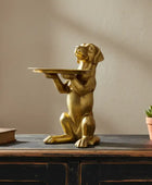 Gold dog statue holding a tray on a wooden surface with a plant and book in the background.