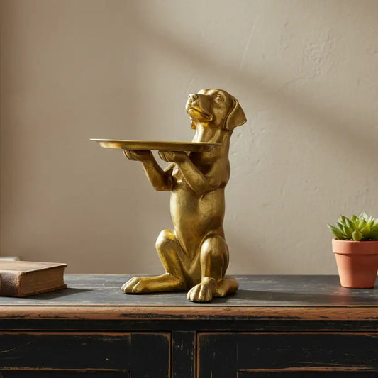 Gold dog statue holding a tray on a wooden surface with a plant and book in the background.