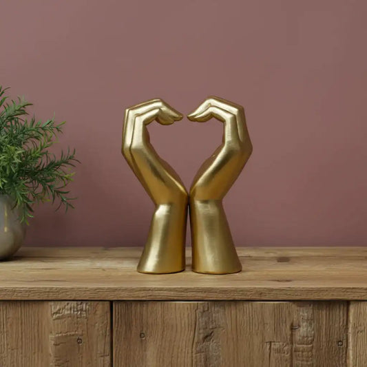 Gold hands sculpture forming a heart shape on a wooden surface with a plant in the background.