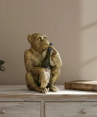 Decorative monkey statue holding a bottle on a wooden surface with a plant and book in the background.