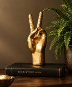 A gold hand sculpture making a peace sign, displayed on a black book titled 'THE KINFOLK HOME' on a wooden table next to a green fern plant.