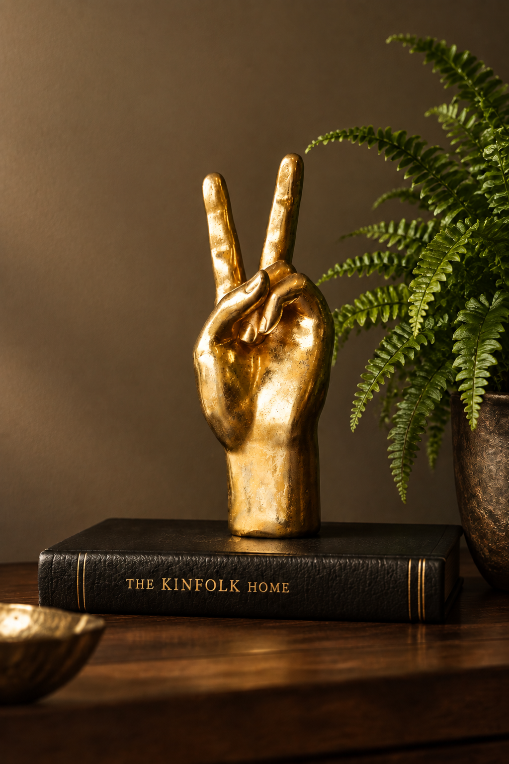 A gold hand sculpture making a peace sign, displayed on a black book titled 'THE KINFOLK HOME' on a wooden table next to a green fern plant.