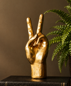 A gold-finished decorative sculpture of a hand making the peace sign, displayed on a black book next to a green potted fern against a warm brown background.