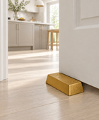 A polished gold door stopper shaped like a gold bar resting on a light wood floor in a doorway, with a modern kitchen visible in the background.