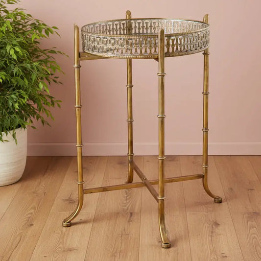 Decorative brass side table with a glass top on a wooden floor against a pink wall.