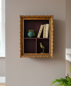 Decorative wall shelf with gold frame containing books, a bird figurine, and a green vase.