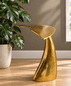 A sculptural modern side table with a textured metallic gold finish, styled in a room next to a potted monstera plant on a light wood floor.