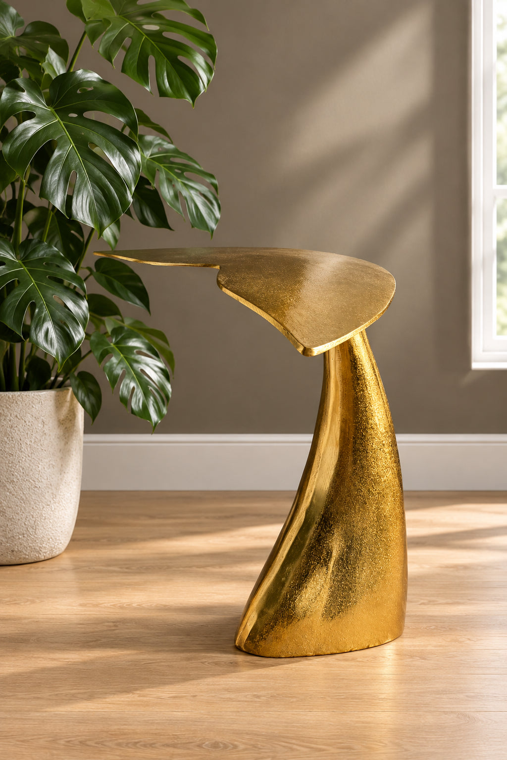 A sculptural modern side table with a textured metallic gold finish, styled in a room next to a potted monstera plant on a light wood floor.