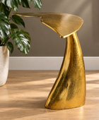 A sculptural modern side table with a metallic gold finish, styled in a room setting next to a potted monstera plant on a light wood floor.