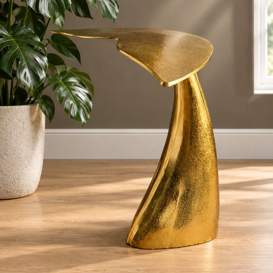 A sculptural modern side table with a metallic gold finish, styled in a room setting next to a potted monstera plant on a light wood floor.