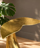 A sculptural leaf-shaped side table in textured gold finish, styled on a wooden floor next to a potted monstera plant against a neutral gray wall.