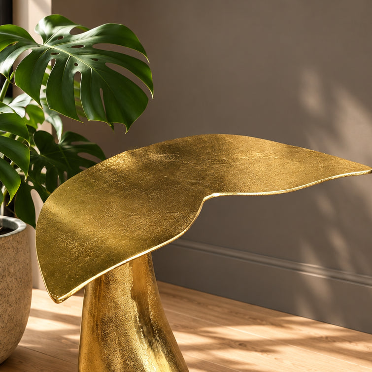 A sculptural leaf-shaped side table in textured gold finish, styled on a wooden floor next to a potted monstera plant against a neutral gray wall.