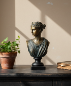 Bronze bust of a woman on a wooden surface with a plant and book in the background
