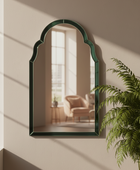 Decorative mirror with a green frame on a beige wall, reflecting a room with furniture and a plant.