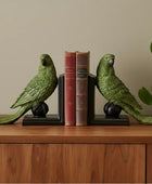 Green parrot bookends on a wooden surface with books between them against a beige wall.