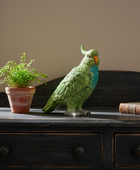 Decorative parrot figurine on a wooden surface with a plant and books in the background.