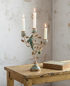 An ornate three-arm candelabra with a gold-toned metal frame, decorative branches, and a green bird figure, shown lit on a distressed wooden table against a textured off-white wall. A vintage book rests beside it.