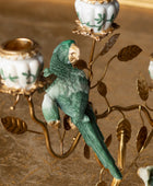 A decorative metal and ceramic candelabra featuring a green parrot perched on gold-toned branches with white candle holders decorated with green floral motifs.