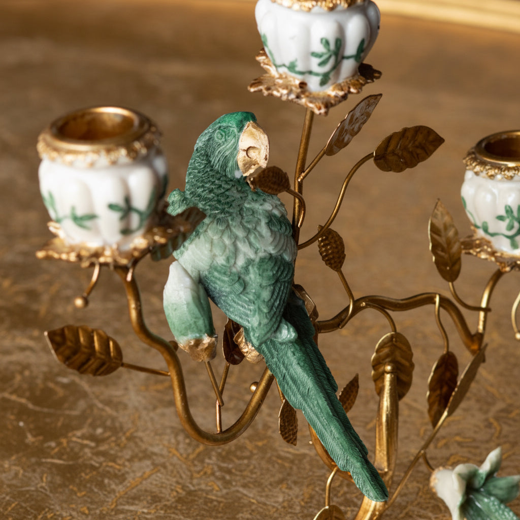A decorative metal and ceramic candelabra featuring a green parrot perched on gold-toned branches with white candle holders decorated with green floral motifs.