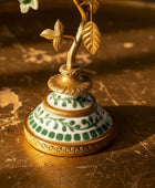 An ornate vintage-style metal and porcelain candlestick holder with a gold-tone branch and leaves, decorated with a white and green porcelain base, set on a rustic wooden surface.