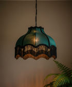 A vintage-style teal glass pendant lamp with black fringe trim, hanging against a warm beige wall with a green fern in the foreground.