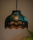 A teal fabric pendant lampshade with black fringe trim and a warm glowing light inside, hanging against a beige wall with a plant leaf visible in the lower right corner.