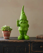 Green gnome figurine on a wooden surface with a potted plant and book in the background.