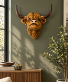 A realistic Highland cow skull wall mount with long curved horns, displayed on a dark olive green wall in a styled room setting with a wooden console table, potted plant, and window.