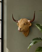 A decorative Highland cow head wall mount with long curved horns displayed on a sage green wall in a styled living room setting with a potted plant and window.