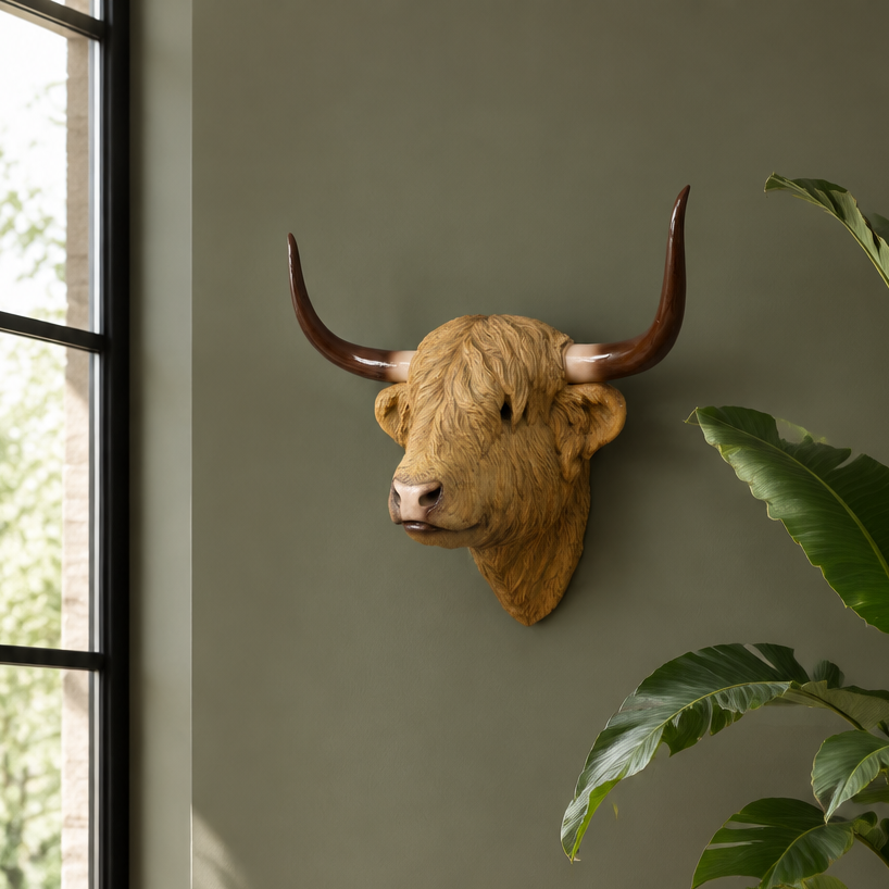 A decorative Highland cow head wall mount with long curved horns displayed on a sage green wall in a styled living room setting with a potted plant and window.