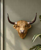 A realistic bull head wall mount with long curved horns, displayed on a muted sage green wall next to a window frame and green houseplant leaves.