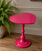 A bright hot pink heart-shaped side table with a turned pedestal base, styled on a rustic wooden floor next to a green potted fern.