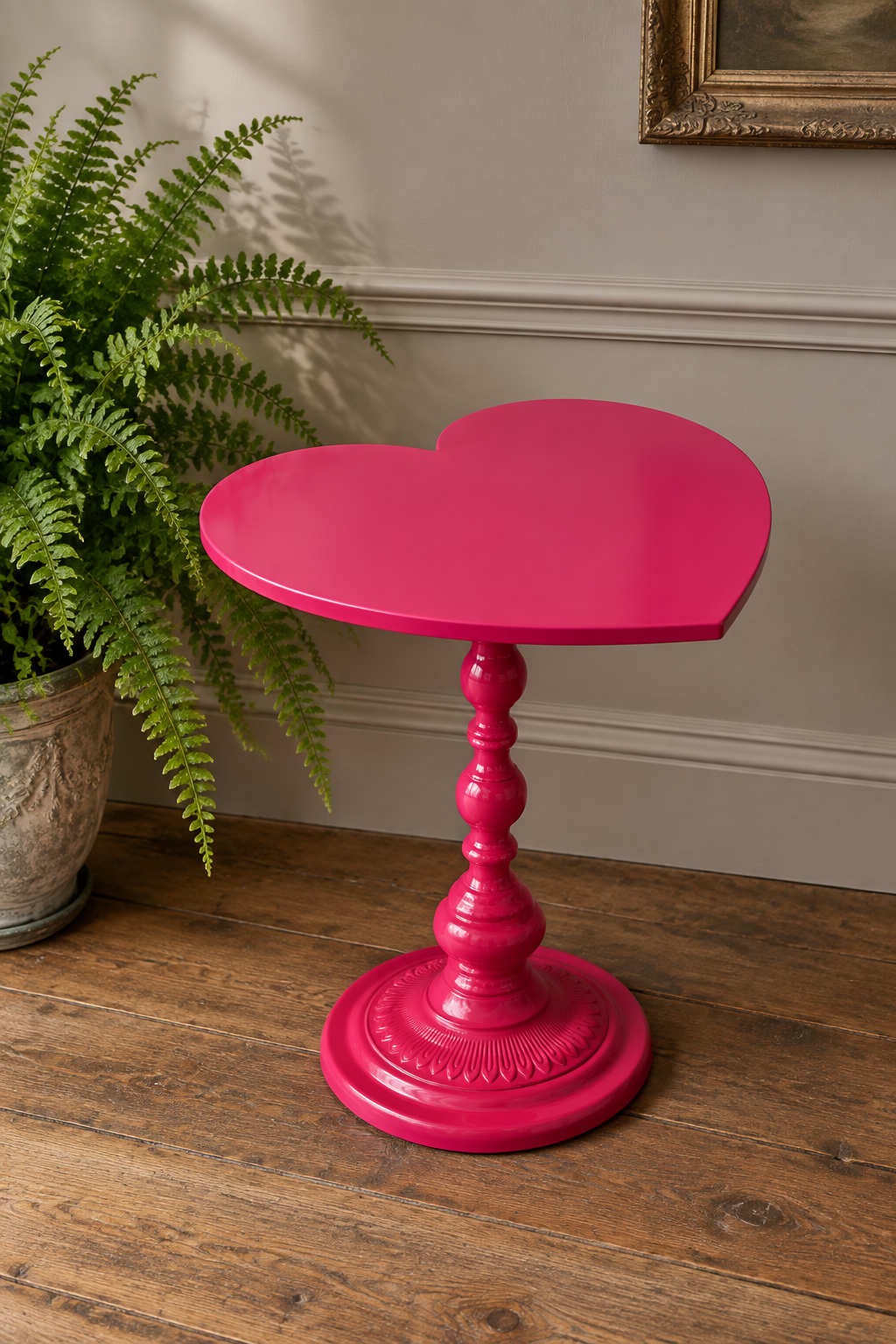 A bright hot pink heart-shaped side table with a turned pedestal base, styled on a rustic wooden floor next to a green potted fern.