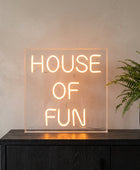 A square acrylic LED neon sign glowing with the words 'HOUSE OF FUN' in a warm orange light, displayed on a dark surface next to two lit candles in holders.