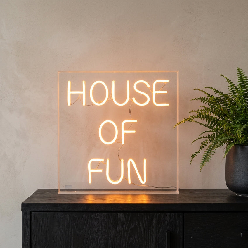 A square acrylic LED neon sign glowing with the words 'HOUSE OF FUN' in a warm orange light, displayed on a dark surface next to two lit candles in holders.