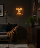 A glowing square neon sign reading 'HOUSE OF FUN' mounted on a dark wall in a styled living room with a sofa, throw blanket, sideboard, framed art, and a potted plant.