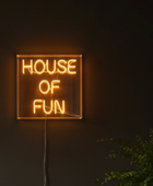 A square neon sign glowing with the words 'HOUSE OF FUN' in warm orange light, mounted on a dark wall with a visible power cord and a small green plant in the lower right corner.