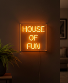 A square neon sign with the words 'HOUSE OF FUN' glowing in warm yellow-orange light, mounted on a dark wall in a dimly lit living room. A potted fern sits on a small table to the left, and a blurred sofa is visible in the background.