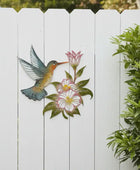 Decorative metal wall art of a hummingbird and flowers on a white picket fence.