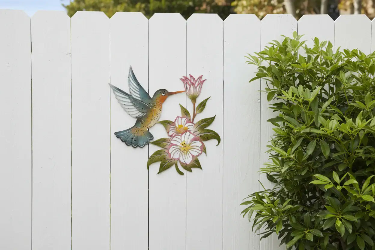 Decorative metal wall art of a hummingbird and flowers on a white picket fence.