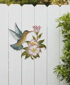 Decorative metal wall art of a hummingbird and flowers on a white wooden fence.