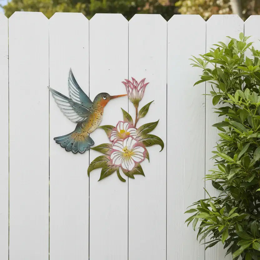 Decorative metal wall art of a hummingbird and flowers on a white wooden fence.