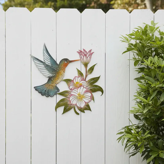 Decorative metal wall art of a hummingbird and flowers on a white wooden fence.