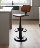A modern kitchen bar stool with a brown leather seat and backrest, set in a bright kitchen with light wood floors and gray cabinets.