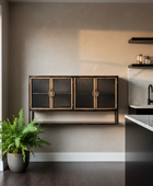 A modern wall-mounted cabinet with three ribbed glass doors in a warm black finish and gold-toned handles, installed in a minimalist kitchen setting with dark wood floors and a potted fern nearby.