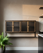 A modern wall-mounted cabinet with three ribbed glass doors and brass-toned handles, shown installed on a beige wall in a styled interior with a potted fern and kitchen elements nearby.