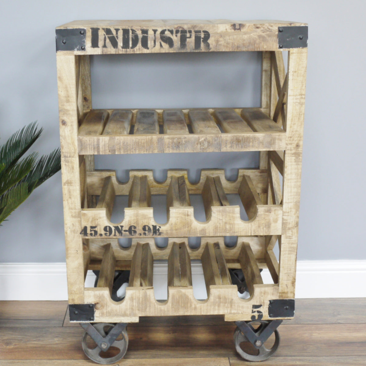 A rustic three-tier wooden rolling shelf cart made from pallet wood, featuring metal corner and wheel brackets, with black stenciled text on a light gray wall and wood floor background.